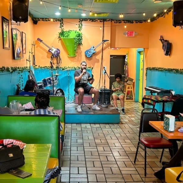 A man speaks on a stage to restaurant patrons seated at green booths and tables.